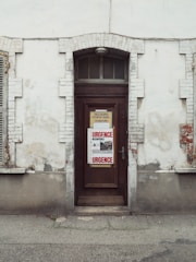 A weathered, brown wooden door set into a brick and plaster wall. The wall is aged with visible cracks and peeling paint. There are two signs on the door, one in the center with 'URGENCE' written in red letters, indicating an emergency, and another above it with French text. The exterior of the building appears old and slightly neglected.