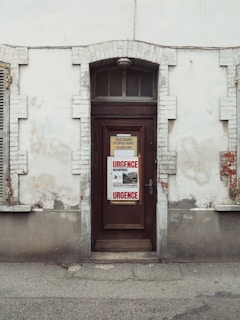 A weathered, brown wooden door set into a brick and plaster wall. The wall is aged with visible cracks and peeling paint. There are two signs on the door, one in the center with 'URGENCE' written in red letters, indicating an emergency, and another above it with French text. The exterior of the building appears old and slightly neglected.