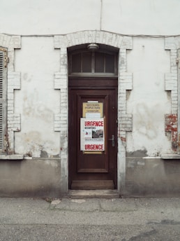 A weathered, brown wooden door set into a brick and plaster wall. The wall is aged with visible cracks and peeling paint. There are two signs on the door, one in the center with 'URGENCE' written in red letters, indicating an emergency, and another above it with French text. The exterior of the building appears old and slightly neglected.