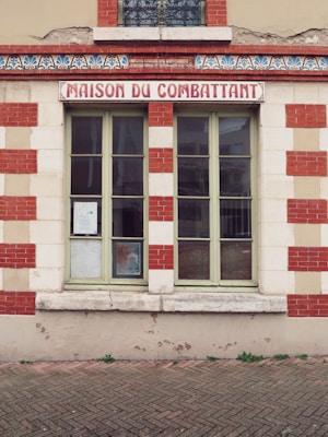 A facade of a building with a central window, framed by red and white bricks, and a sign above reading 'Maison du Combattant'. There is decorative molding underneath the sign and slight wear visible on the building's surface.