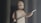 A stylized animated character with a simple facial expression stands holding a spear in one hand. The background features large, shadowy columns, creating an ancient or mystical atmosphere. The character has a bald head and a beard, wearing a basic one-shoulder garment.