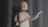 A stylized animated character with a simple facial expression stands holding a spear in one hand. The background features large, shadowy columns, creating an ancient or mystical atmosphere. The character has a bald head and a beard, wearing a basic one-shoulder garment.