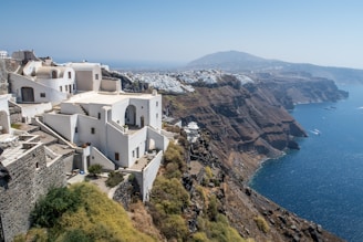Whitewashed buildings with blue domes and arches cascade down a steep cliffside overlooking a deep blue sea. The landscape includes rugged brown cliffs, while the town stretches along the cliff edge in the distance. The architecture and natural beauty combine harmoniously, showcasing vibrant sea and sky.