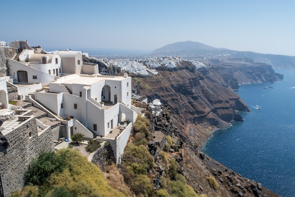 Whitewashed buildings with blue domes and arches cascade down a steep cliffside overlooking a deep blue sea. The landscape includes rugged brown cliffs, while the town stretches along the cliff edge in the distance. The architecture and natural beauty combine harmoniously, showcasing vibrant sea and sky.
