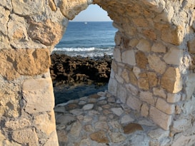 A stone archway constructed from irregularly shaped rocks stands beside an expansive seascape. The ocean waves gently crash onto a rugged rocky shoreline, and the blue water stretches towards the horizon, where a small boat is visible.