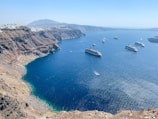 A stunning coastal landscape featuring a deep blue sea with several large cruise ships and small boats traversing the water. The coastline comprises rugged cliffs with earthy tones and sparse vegetation. In the distance, mountains rise against a clear blue sky, and clusters of white buildings are dotted along the top of the cliffs.