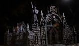 An intricate gothic structure features archways adorned with skulls. The dark, moody lighting accentuates the detailed architecture, including pointed spires and ornate stonework. A statue holding a weapon or shield stands atop the structure, adding to the eerie, mysterious atmosphere.