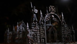An intricate gothic structure features archways adorned with skulls. The dark, moody lighting accentuates the detailed architecture, including pointed spires and ornate stonework. A statue holding a weapon or shield stands atop the structure, adding to the eerie, mysterious atmosphere.