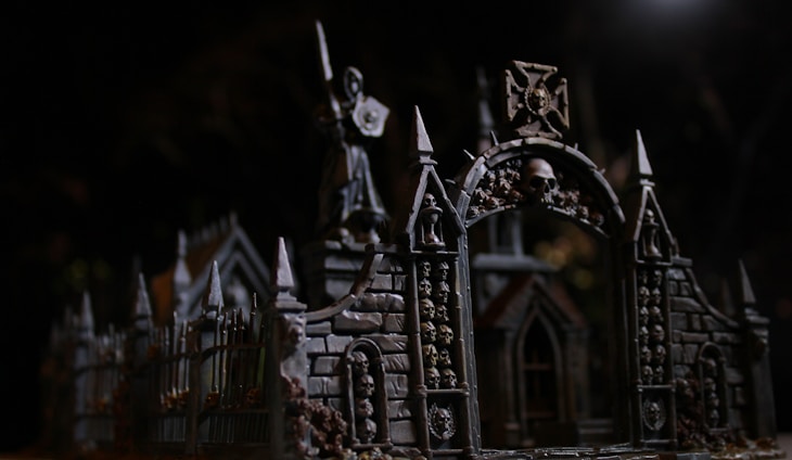 An intricate gothic structure features archways adorned with skulls. The dark, moody lighting accentuates the detailed architecture, including pointed spires and ornate stonework. A statue holding a weapon or shield stands atop the structure, adding to the eerie, mysterious atmosphere.