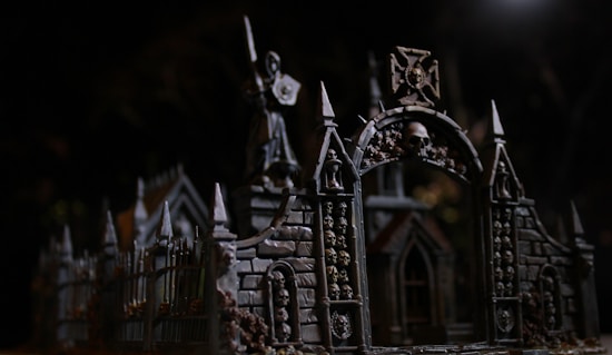 An intricate gothic structure features archways adorned with skulls. The dark, moody lighting accentuates the detailed architecture, including pointed spires and ornate stonework. A statue holding a weapon or shield stands atop the structure, adding to the eerie, mysterious atmosphere.
