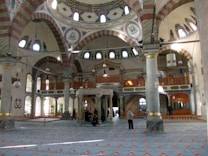 A spacious and ornately decorated interior of a mosque featuring large marble columns, high arched ceilings, and intricate designs with Arabic calligraphy. The space is illuminated by natural light coming through numerous windows. A few people are present, some standing and some seated on the decorative carpet that covers the floor.