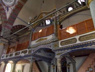 An intricately designed interior with ornate wooden and column details, featuring decorative patterns and arches. The architecture includes painted motifs, geometric designs, and carved elements.