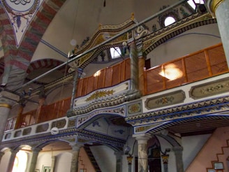 An intricately designed interior with ornate wooden and column details, featuring decorative patterns and arches. The architecture includes painted motifs, geometric designs, and carved elements.