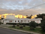 A residential area with a single-story house in the foreground and multi-story buildings in the background. The house has white walls with light pink accents and a small garden with trimmed bushes and a neat lawn. The sky is cloudy with parts of the clouds illuminated by the setting sun, casting a warm glow on the buildings in the background.