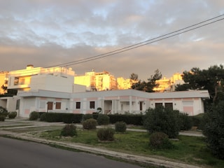 A residential area with a single-story house in the foreground and multi-story buildings in the background. The house has white walls with light pink accents and a small garden with trimmed bushes and a neat lawn. The sky is cloudy with parts of the clouds illuminated by the setting sun, casting a warm glow on the buildings in the background.