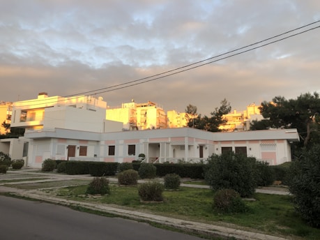 A residential area with a single-story house in the foreground and multi-story buildings in the background. The house has white walls with light pink accents and a small garden with trimmed bushes and a neat lawn. The sky is cloudy with parts of the clouds illuminated by the setting sun, casting a warm glow on the buildings in the background.