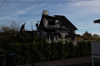 A partially burned house with visible charred walls and a damaged roof surrounded by green bushes. The sky is clear with some clouds. There is a fence in front of the house.