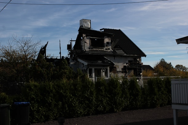 A partially burned house with visible charred walls and a damaged roof surrounded by green bushes. The sky is clear with some clouds. There is a fence in front of the house.