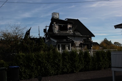 A partially burned house with visible charred walls and a damaged roof surrounded by green bushes. The sky is clear with some clouds. There is a fence in front of the house.