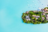 An aerial view of a small peninsula extending into vibrant turquoise water, featuring a cluster of houses surrounded by lush green vegetation. Several small boats are moored nearby, adding a peaceful touch to the scene.