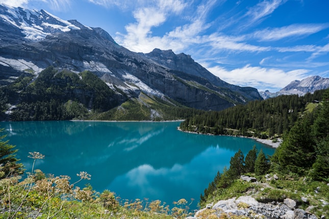 A serene landscape featuring a turquoise lake surrounded by dense evergreen forests and towering snow-capped mountains under a clear blue sky with wispy clouds.