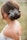 A woman with a delicate floral hair accessory is seen from the back. Her hair is styled in an elegant updo, and she is wearing an off-the-shoulder lace dress. A tattoo is visible on her upper back, and the background is softly blurred, suggesting an outdoor setting.