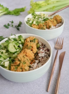 Two bowls filled with a mix of rice, chopped cucumber, leafy greens, and falafel balls sit on a light gray surface. Fresh green celery and parsley are visible in the background. A pair of bronze-colored fork and spoon are placed next to the bowls.