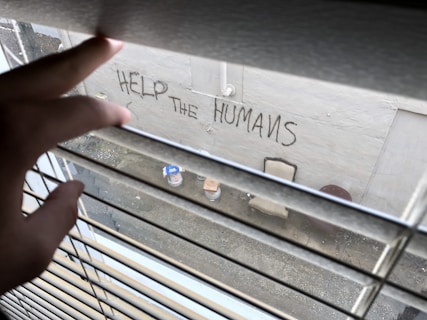 A hand is seen partially opening Venetian blinds, looking out onto a scene where 'HELP THE HUMANS' is written on a wall. Below, there are a couple of objects, including a chair and a container.