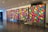 A series of four large panels display a vibrant, geometric pattern composed of multicolored rectangles. The artwork is installed in a modern, spacious gallery with a reflective marble floor and recessed lighting. Each panel features a varied arrangement of colorful shapes, creating a dynamic visual rhythm.