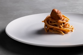 A small serving of spaghetti topped with a single meatball is placed neatly in the center of a large, plain white plate. The setting is minimalistic with a soft-focus gray background.