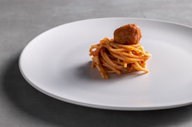 A minimalist serving consisting of a small portion of spaghetti neatly twisted and topped with a single meatball, presented on a large white plate against a neutral gray background. The spaghetti is lightly coated with red sauce, giving a hint of flavor and contrast.