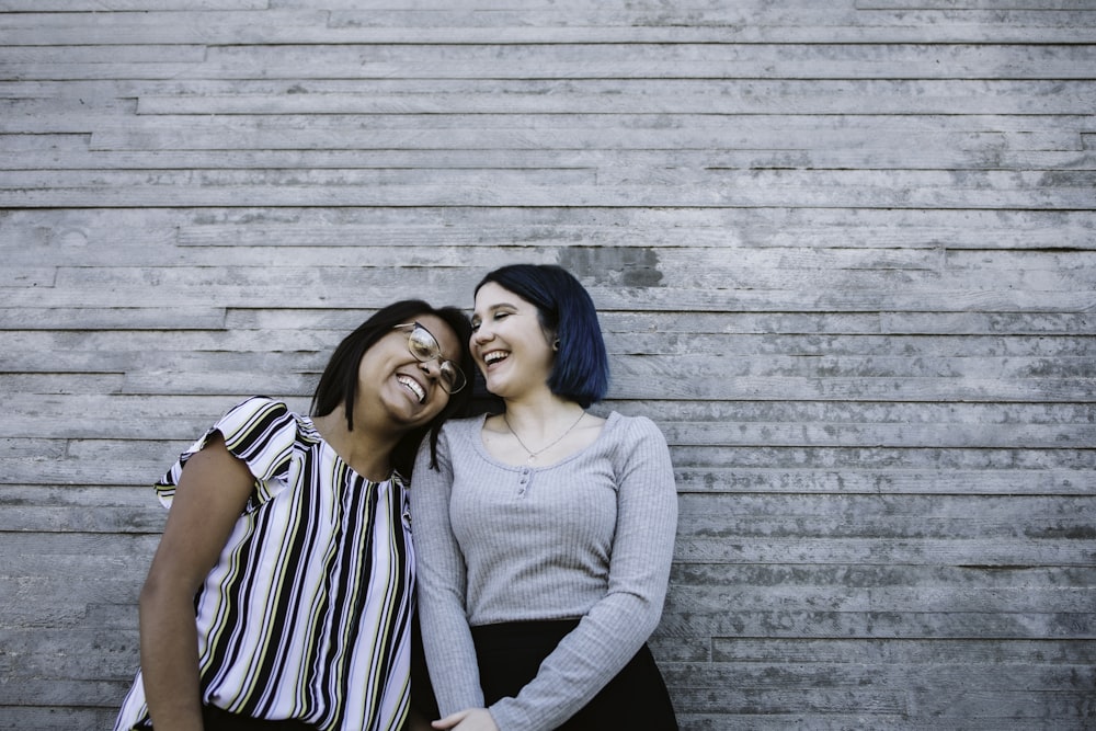 2 women smiling and standing beside gray wooden wall