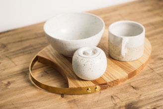 A set of ceramic tableware including a bowl, a mug, and a container with a lid rests on a wooden board with a metal handle. The ceramics are speckled with a subtle dotted pattern, and the setup is placed on a wooden surface.