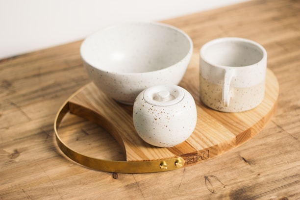 A set of ceramic tableware including a bowl, a mug, and a container with a lid rests on a wooden board with a metal handle. The ceramics are speckled with a subtle dotted pattern, and the setup is placed on a wooden surface.