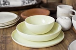 A collection of tableware including a stack of light-colored bowls and plates on a wooden surface, surrounded by additional plates and a speckled ceramic mug with a small teapot.