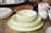 A collection of tableware including a stack of light-colored bowls and plates on a wooden surface, surrounded by additional plates and a speckled ceramic mug with a small teapot.