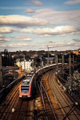 A modern passenger train navigates through a series of curved railway tracks in an urban setting. The train is red and white, and several carriages can be seen extending into the distance. Overhead, a partly cloudy sky casts shadows on the landscape. In the background, a crane and various buildings are visible, suggesting ongoing construction or infrastructure.