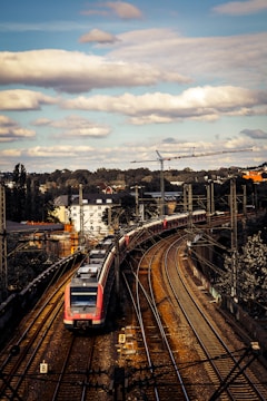 A modern passenger train navigates through a series of curved railway tracks in an urban setting. The train is red and white, and several carriages can be seen extending into the distance. Overhead, a partly cloudy sky casts shadows on the landscape. In the background, a crane and various buildings are visible, suggesting ongoing construction or infrastructure.