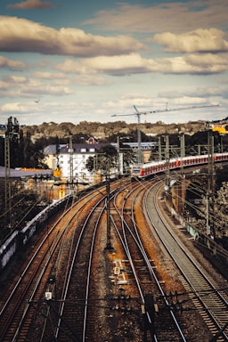 A network of train tracks curving into the distance with a red train on the bridge. Overhead power lines and signals are visible, while a large crane looms in the background. Buildings with red rooftops and lush greenery can be seen under a partly cloudy sky.