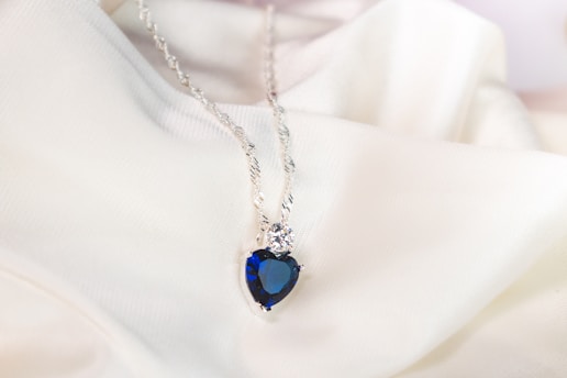 A heart-shaped blue gemstone pendant is suspended from a delicate silver chain, resting on a soft, white fabric background.
