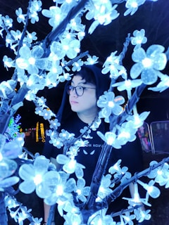 A person wearing glasses and a hood is surrounded by illuminated floral decorations. The white and blue lights from the flower lights create a glowing effect, contrasting with the darkness of the background.