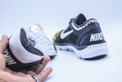 A pair of black and white athletic shoes are displayed. One shoe is held in a hand showing the sole, which has a textured, waffle-like pattern. The other shoe rests on its side, highlighting the brand logo on the heel.