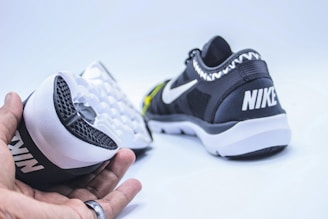 A pair of black and white athletic shoes are displayed. One shoe is held in a hand showing the sole, which has a textured, waffle-like pattern. The other shoe rests on its side, highlighting the brand logo on the heel.