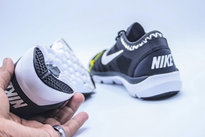 A pair of black and white athletic shoes are displayed. One shoe is held in a hand showing the sole, which has a textured, waffle-like pattern. The other shoe rests on its side, highlighting the brand logo on the heel.
