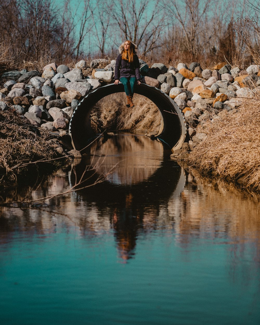 man in blue jacket and blue denim jeans standing on brown rock near body of water