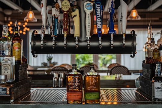 A bar setup with a variety of beer taps hanging from a black metal pipe and several liquor bottles placed on the counter. Two bottles of Bulleit whiskey are prominently displayed in the center. The background features bar stools and soft lighting from hanging pendant lamps.
