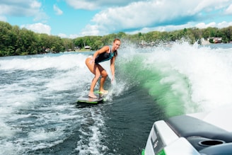 A person is surfing on a wakeboard behind a boat, navigating through the waves with skill. The scene takes place on a lake surrounded by lush green trees and a partly cloudy sky. The wake of the boat creates dynamic, foamy waves, contrasting with the calmness of the surrounding landscape.