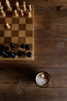 A wooden chessboard with a few wooden and black chess pieces is set on a rustic wooden table. To the side of the chessboard, there is a cup of coffee with artful latte art on the surface, showing a heart-shaped pattern.
