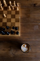 A wooden chessboard with a few wooden and black chess pieces is set on a rustic wooden table. To the side of the chessboard, there is a cup of coffee with artful latte art on the surface, showing a heart-shaped pattern.