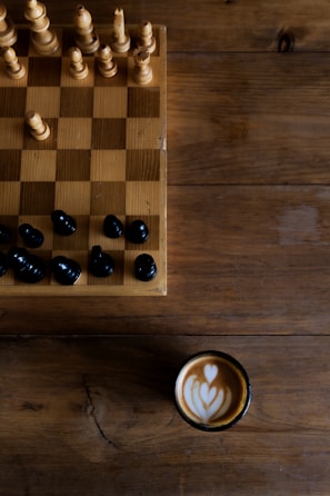 A wooden chessboard with a few wooden and black chess pieces is set on a rustic wooden table. To the side of the chessboard, there is a cup of coffee with artful latte art on the surface, showing a heart-shaped pattern.
